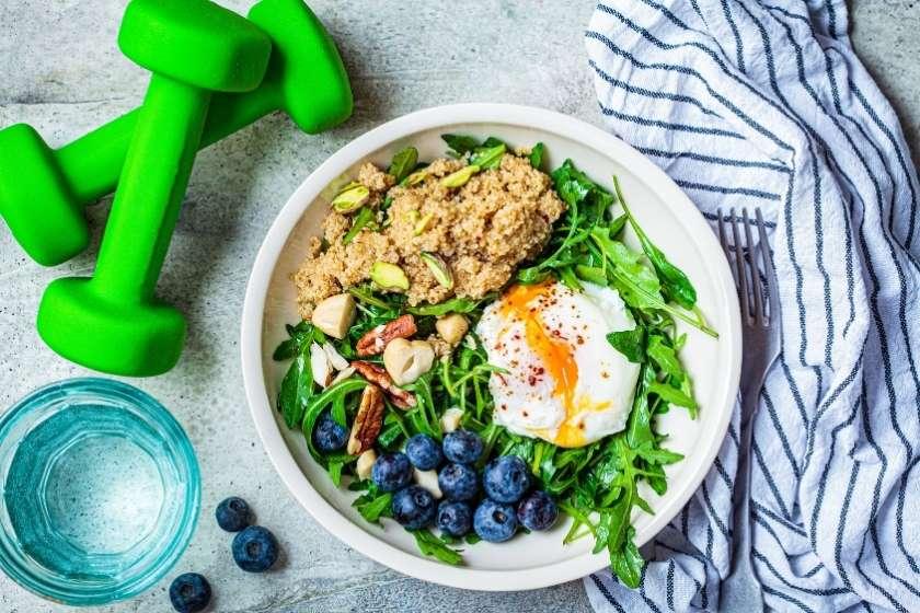 breakfast bowl with poached egg, quinoa, nuts, blueberries, and arugula, next to green dumbbells and a glass of water